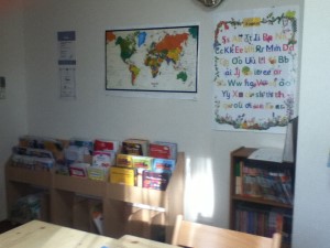 壁にはJolly Phonicsのレターサウンドのポスター、そして世界地図を貼っています。下の方に見えるのがミニライブラリ。好きな本を選んで、借りて帰ってください。